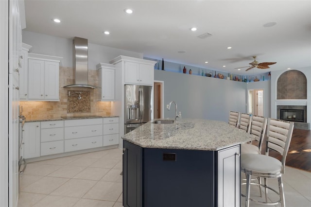 kitchen with a sink, backsplash, stainless steel fridge, a fireplace, and wall chimney range hood