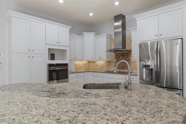 kitchen with backsplash, wall chimney range hood, light stone counters, appliances with stainless steel finishes, and white cabinets