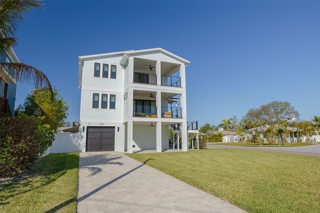 coastal inspired home with a front yard, decorative driveway, a balcony, an attached garage, and a ceiling fan