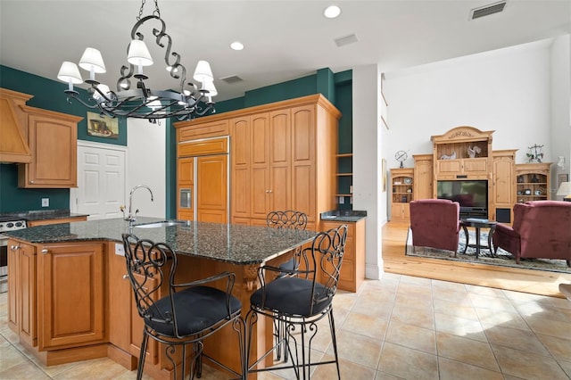 kitchen featuring visible vents, a center island with sink, dark stone counters, paneled fridge, and a sink