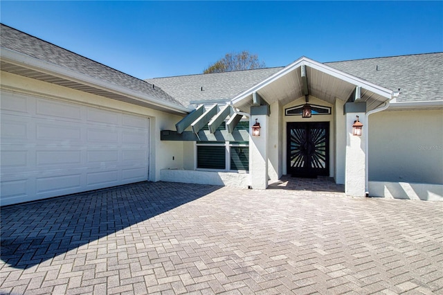 view of exterior entry with stucco siding, decorative driveway, a garage, and roof with shingles