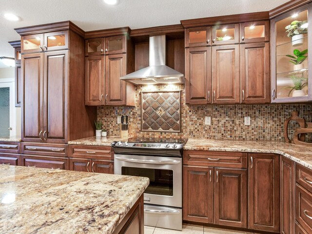 kitchen featuring backsplash, glass insert cabinets, wall chimney range hood, light stone counters, and electric range