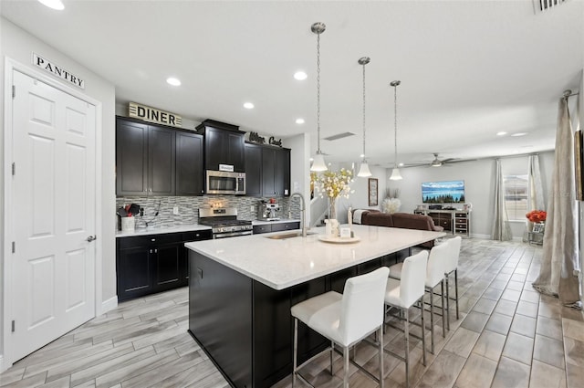 kitchen with a center island with sink, a sink, decorative backsplash, stainless steel appliances, and open floor plan