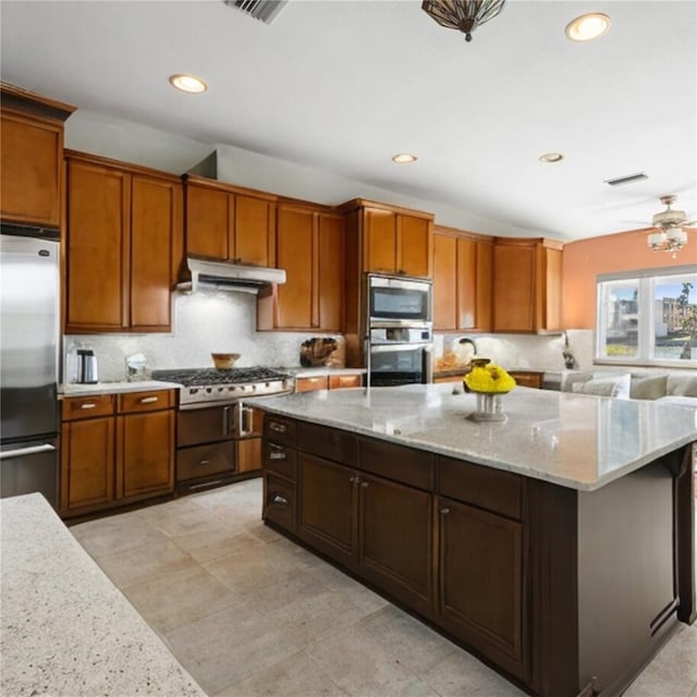 kitchen with under cabinet range hood, light stone counters, a kitchen island, appliances with stainless steel finishes, and decorative backsplash