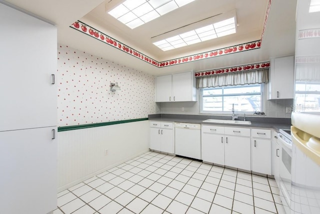 kitchen with white appliances, wallpapered walls, a sink, wainscoting, and white cabinetry