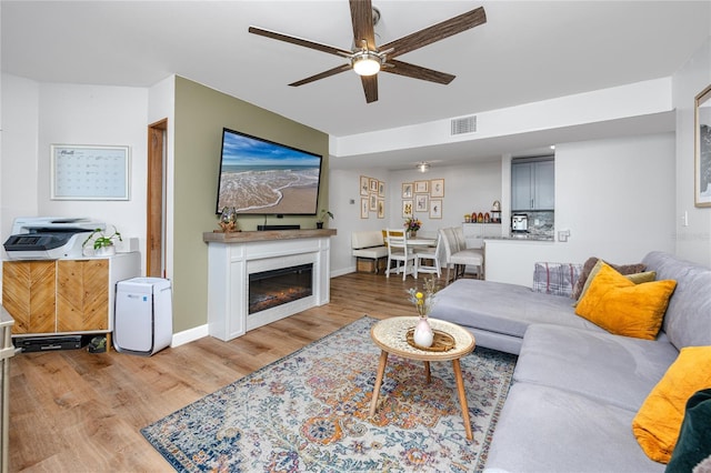 living area with visible vents, a glass covered fireplace, light wood-style floors, baseboards, and ceiling fan