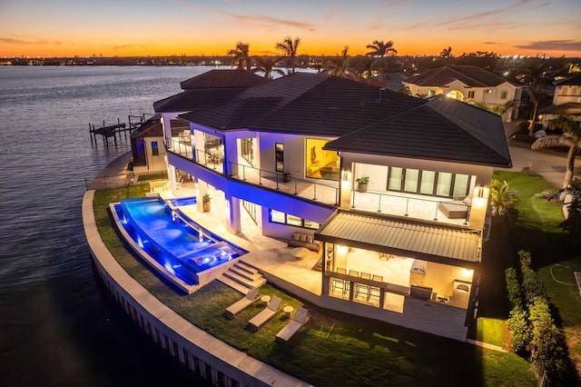 back of property at dusk featuring an infinity pool, a patio area, a water view, and stucco siding