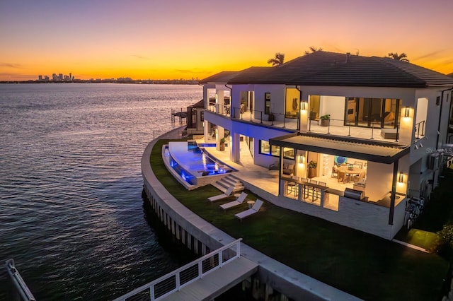 rear view of house with a balcony, a water view, a tile roof, a patio area, and an infinity pool