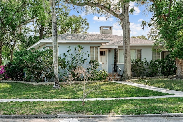 view of front facade with stucco siding, roof with shingles, a chimney, and a front yard