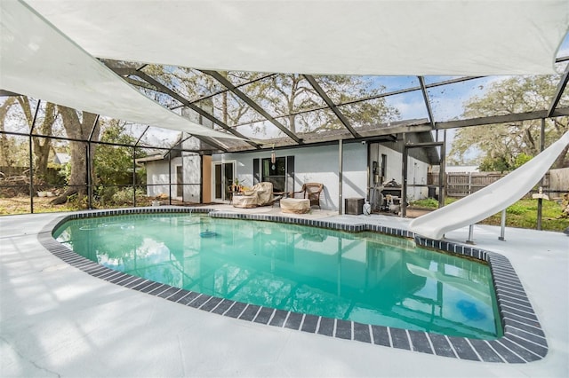 pool with a patio area, glass enclosure, a water slide, and fence