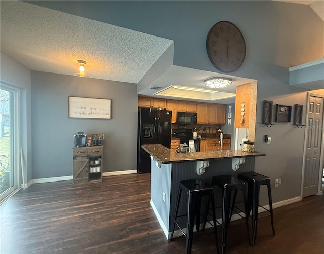 kitchen featuring a peninsula, brown cabinetry, black appliances, a raised ceiling, and dark wood-style flooring