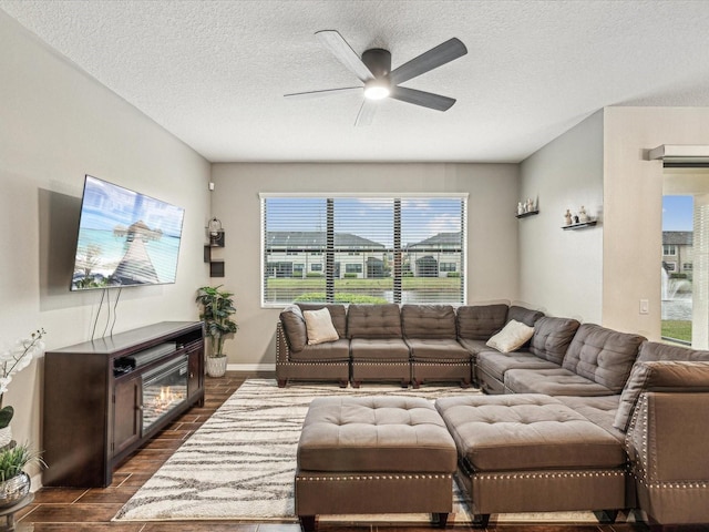 living room with baseboards, ceiling fan, dark wood-type flooring, a textured ceiling, and a glass covered fireplace