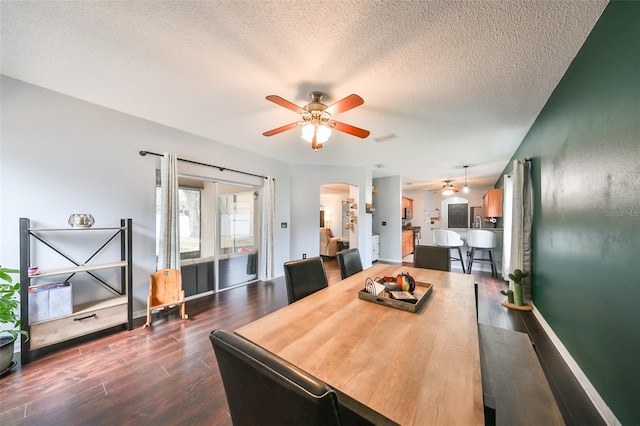 dining room with dark wood-type flooring, a textured ceiling, arched walkways, baseboards, and ceiling fan