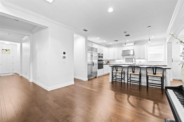 kitchen featuring under cabinet range hood, ornamental molding, a kitchen bar, and stainless steel built in refrigerator