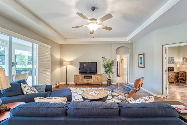 living area featuring wood finished floors, baseboards, a tray ceiling, arched walkways, and ceiling fan