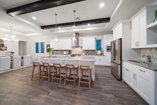 kitchen featuring an island with sink, appliances with stainless steel finishes, wall chimney exhaust hood, white cabinets, and a raised ceiling