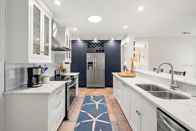 kitchen with light stone counters, a sink, appliances with stainless steel finishes, wall chimney exhaust hood, and backsplash