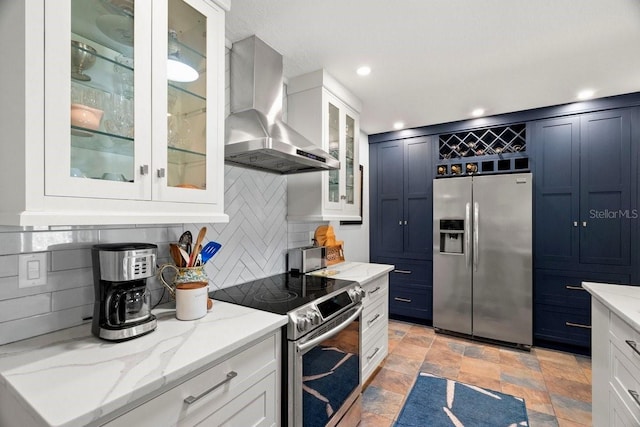 kitchen with light stone counters, stone finish flooring, appliances with stainless steel finishes, wall chimney range hood, and decorative backsplash