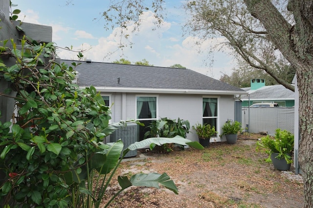 exterior space with cooling unit, roof with shingles, concrete block siding, and fence