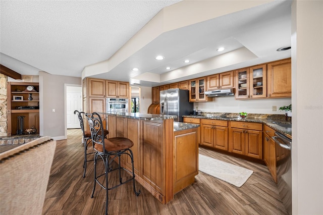 kitchen featuring brown cabinetry, dark wood finished floors, stainless steel appliances, under cabinet range hood, and a center island