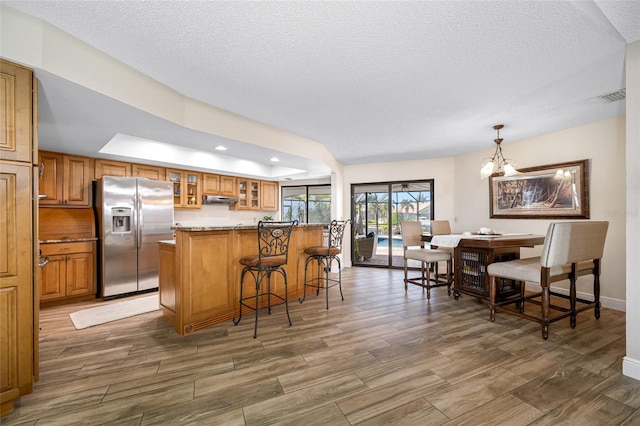 kitchen with under cabinet range hood, wood finished floors, a breakfast bar area, brown cabinetry, and stainless steel fridge with ice dispenser