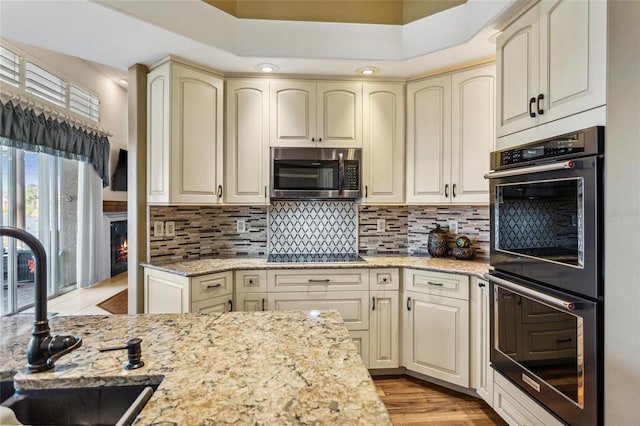 kitchen with a sink, cream cabinetry, stainless steel microwave, black electric stovetop, and multiple ovens