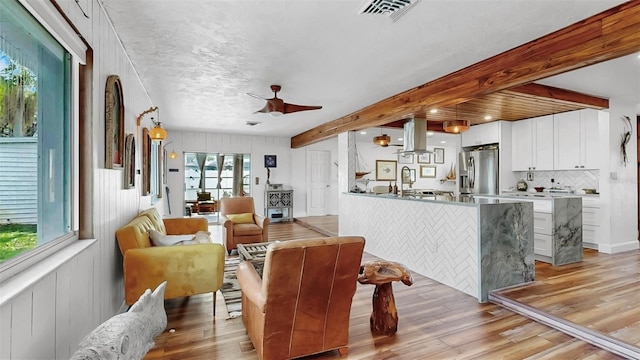 living area featuring light wood-type flooring, beamed ceiling, visible vents, and a ceiling fan