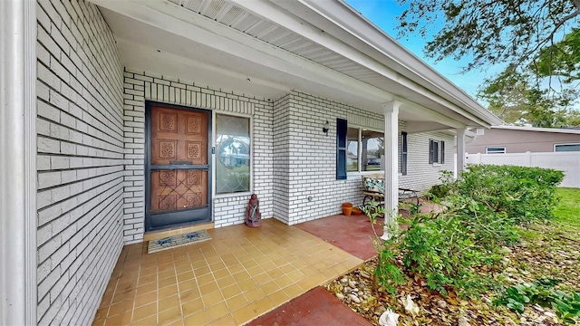 view of exterior entry with brick siding, covered porch, and fence