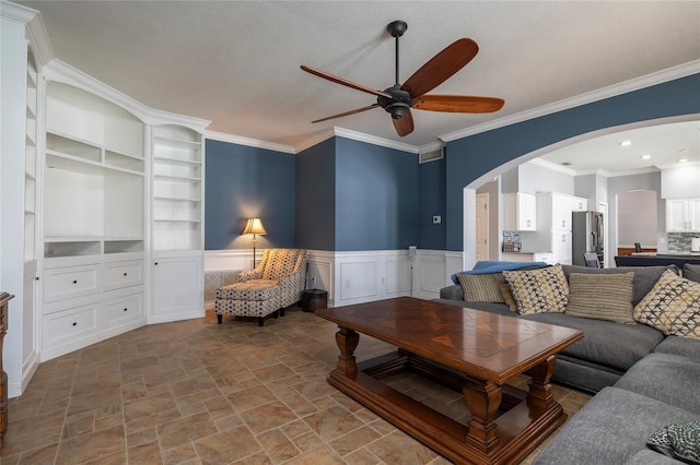 living room with built in shelves, ornamental molding, arched walkways, wainscoting, and ceiling fan