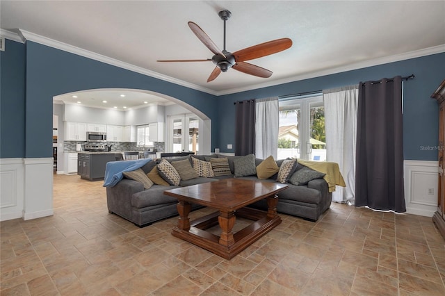 living area featuring a ceiling fan, arched walkways, a wainscoted wall, and ornamental molding