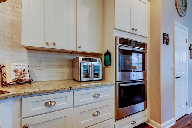 kitchen featuring light stone counters, backsplash, double oven, white cabinets, and baseboards