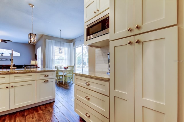 kitchen with stainless steel microwave, backsplash, dark wood-type flooring, light stone countertops, and decorative light fixtures