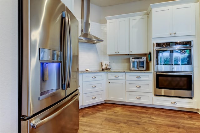 kitchen featuring light stone counters, wall chimney range hood, appliances with stainless steel finishes, white cabinets, and light wood finished floors