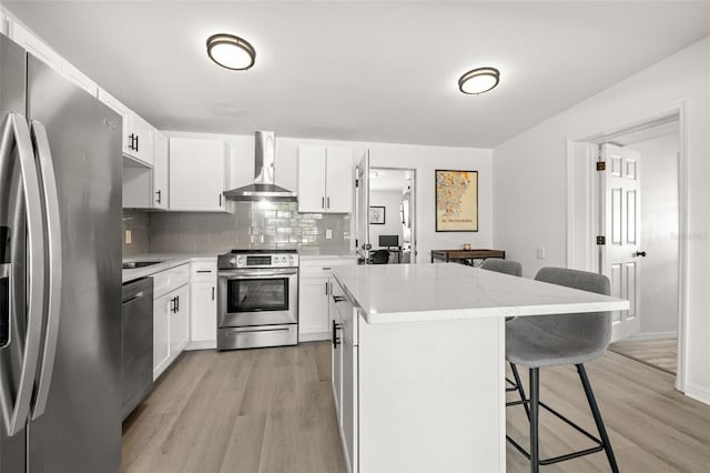 kitchen with wall chimney range hood, a kitchen island, light wood-style floors, appliances with stainless steel finishes, and a kitchen breakfast bar