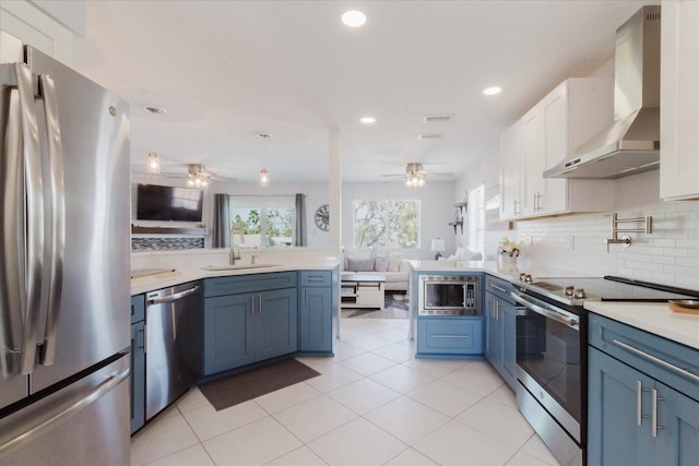 kitchen featuring wall chimney range hood, open floor plan, appliances with stainless steel finishes, a peninsula, and a ceiling fan