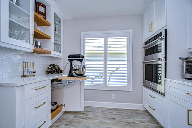 kitchen featuring light wood-type flooring, backsplash, stainless steel double oven, white cabinets, and glass insert cabinets