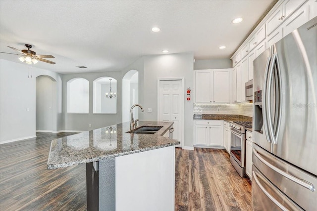 kitchen with a sink, dark wood finished floors, appliances with stainless steel finishes, arched walkways, and a kitchen island with sink