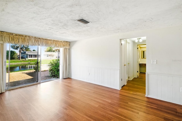 unfurnished room featuring visible vents, a water view, a textured ceiling, wood finished floors, and wainscoting