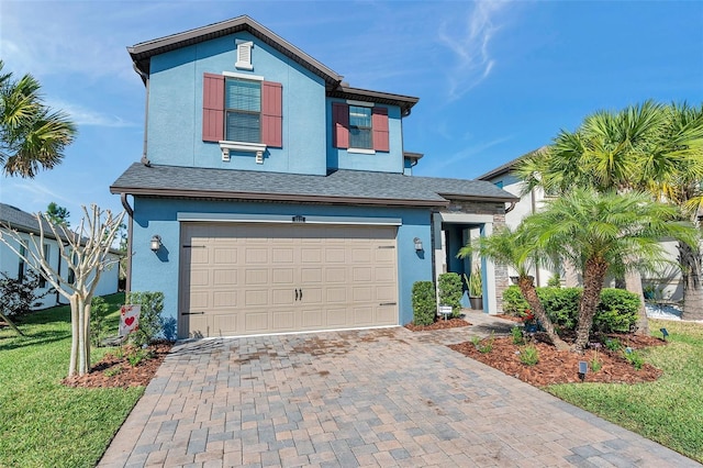 traditional-style home featuring stucco siding, decorative driveway, roof with shingles, a front yard, and a garage