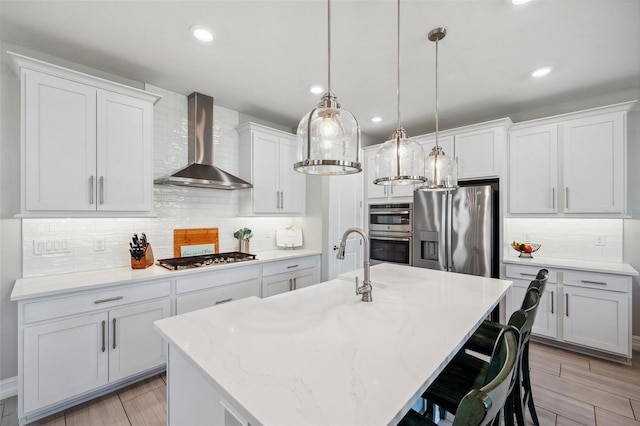 kitchen with an island with sink, decorative light fixtures, stainless steel appliances, white cabinets, and wall chimney range hood