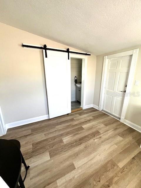 unfurnished bedroom featuring wood finished floors, baseboards, vaulted ceiling, a textured ceiling, and a barn door