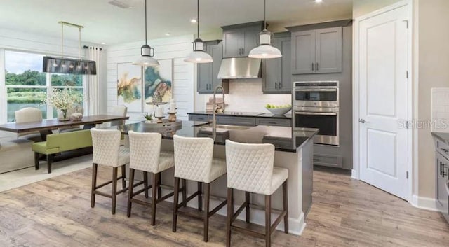 kitchen with stainless steel double oven, an island with sink, gray cabinets, extractor fan, and dark countertops