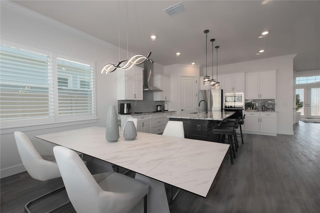 kitchen with a center island with sink, stainless steel appliances, white cabinets, a kitchen bar, and wall chimney exhaust hood