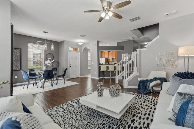 living room featuring visible vents, stairway, ceiling fan with notable chandelier, wood finished floors, and arched walkways