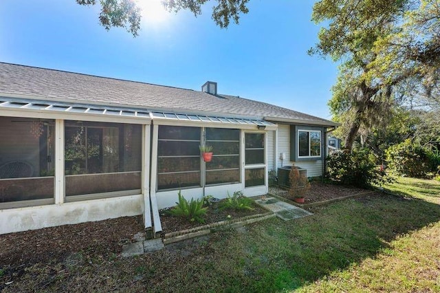 rear view of property featuring a shingled roof, central AC, a lawn, a chimney, and a sunroom