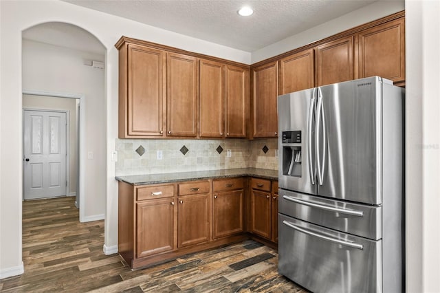 kitchen featuring backsplash, dark stone counters, arched walkways, brown cabinetry, and stainless steel fridge with ice dispenser