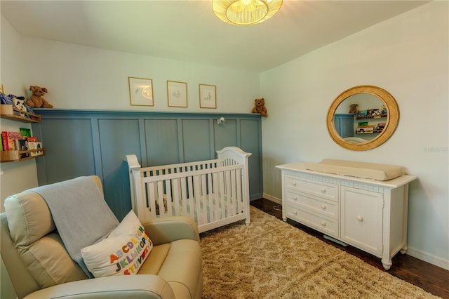 bedroom with baseboards, a crib, dark wood-style floors, and a decorative wall