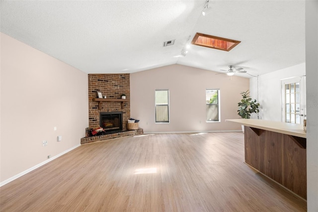 unfurnished living room featuring visible vents, a brick fireplace, light wood-type flooring, vaulted ceiling with skylight, and a ceiling fan