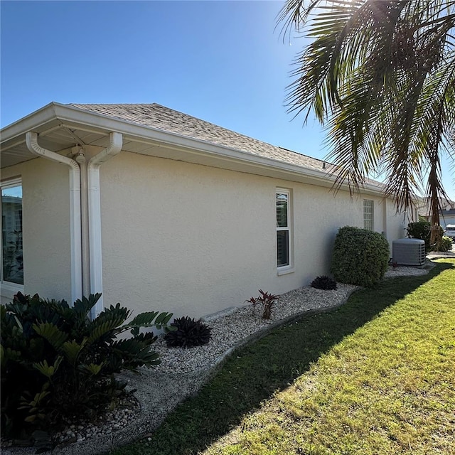 view of side of property featuring a shingled roof, a yard, central AC unit, and stucco siding