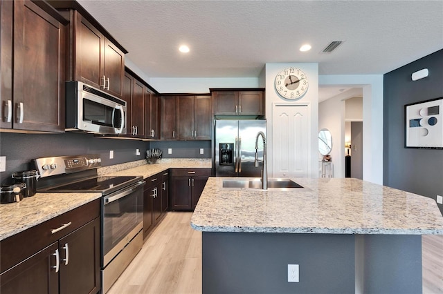 kitchen with dark brown cabinets, a center island with sink, light wood-style flooring, stainless steel appliances, and a sink